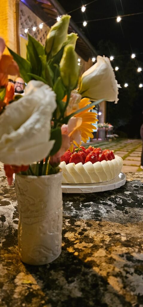 Torta artigianale senza glutine con crema alla vaniglia, composta di fragole e decorazione di fragole fresche e fiori di sambuco, esposta su un’alzatina in ceramica bianca.