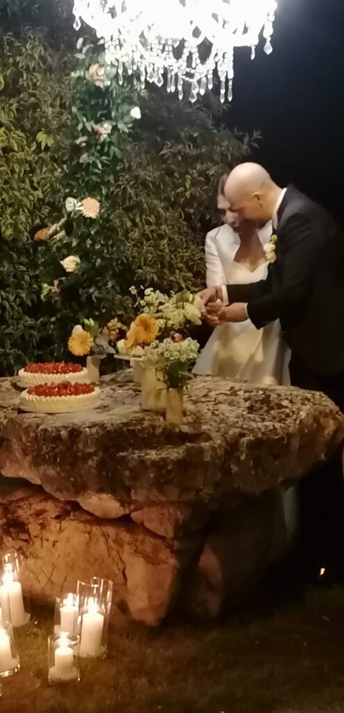 Coppia di sposi ritratti mentre sono al taglio della Torta Mani al loro matrimonio. La scena è ambientata all'esterno e l'atmosfera è elegante.