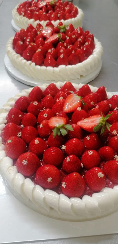 tris di Torta artigianale senza glutine con crema alla vaniglia, composta di fragole e decorazione di fragole fresche e fiori di sambuco, esposta su un’alzatina in ceramica bianca.