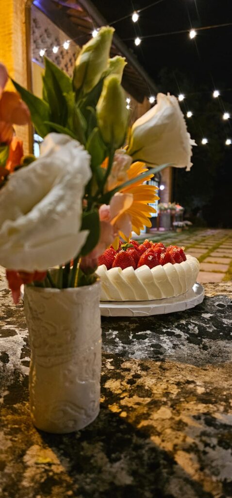 Torta artigianale senza glutine con crema alla vaniglia, composta di fragole e decorazione di fragole fresche e fiori di sambuco, esposta su un’alzatina in ceramica bianca.